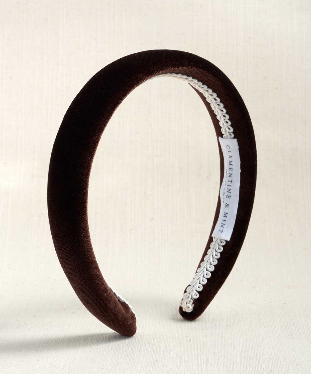 A dark brown velvet headband with a white lace trim and a white brand label, displayed on a light beige textured background.