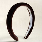 A dark brown velvet headband with a white lace trim and a white brand label, displayed on a light beige textured background.