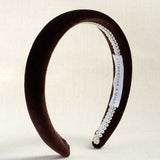 A dark brown velvet headband with a white lace trim and a white brand label, displayed on a light beige textured background.
