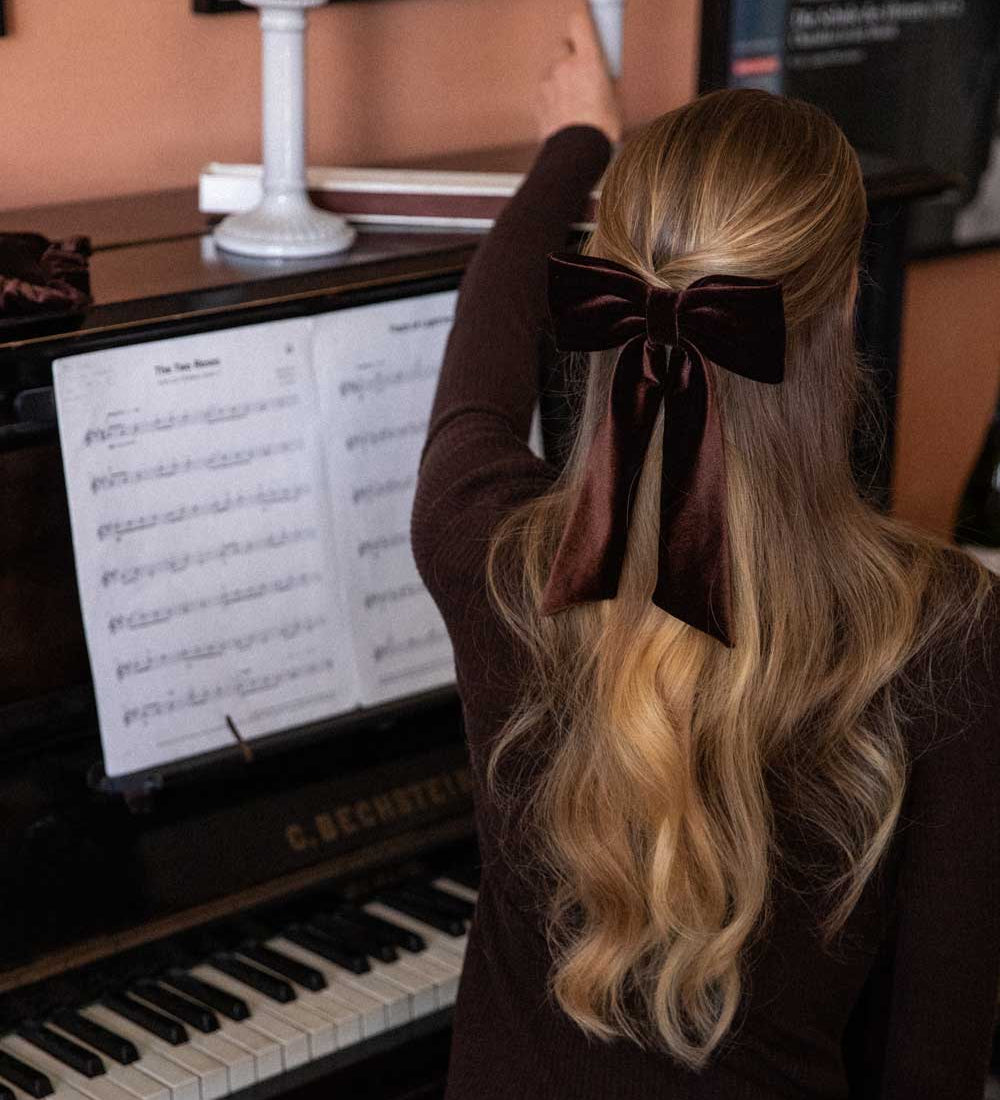Person with long blonde hair and a brown velvet bow sitting at a piano.