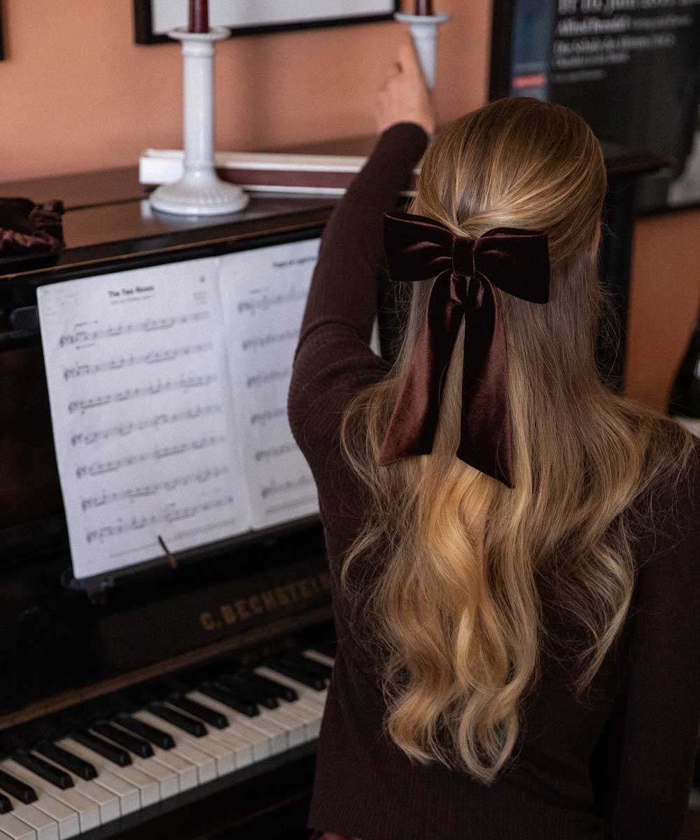 Person with long blonde hair and a brown velvet bow sitting at a piano.