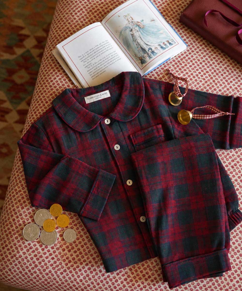 Childrens burgundy tartan pyjamas laid out on fabric with bells and a book 