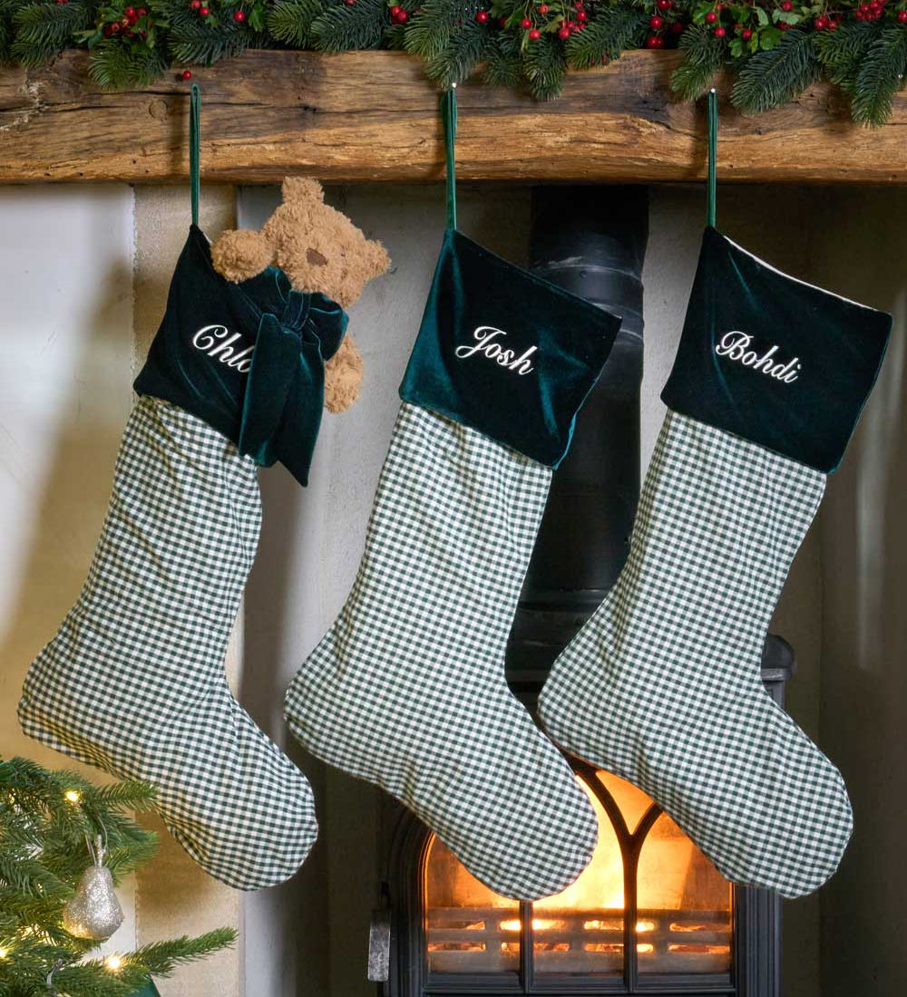 Three personalized Christmas stockings with green and white gingham bodies and dark green velvet cuffs hang from a wooden mantel. The cuffs are embroidered with the names 'Chloé', 'Josh', and 'Boshidi' in white script.