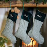 Three personalized Christmas stockings with green and white gingham bodies and dark green velvet cuffs hang from a wooden mantel. The cuffs are embroidered with the names 'Chloé', 'Josh', and 'Boshidi' in white script.