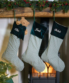 Three personalized Christmas stockings with green and white gingham bodies and dark green velvet cuffs hang from a wooden mantel. The cuffs are embroidered with the names 'Chloé', 'Josh', and 'Boshidi' in white script.
