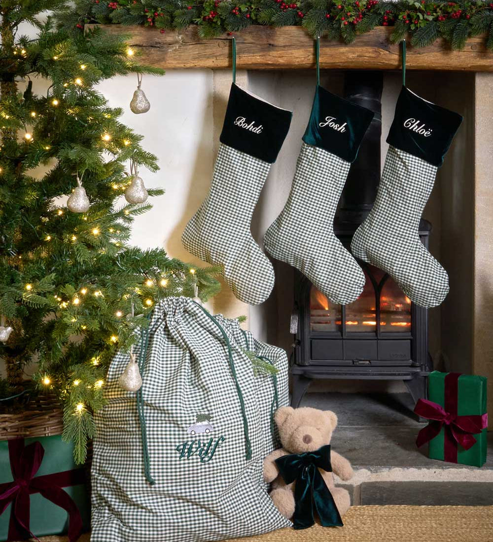 Three personalised Christmas stockings with green and white gingham check patterns and forest green velvet cuffs hang on a rustic wooden mantel above a burning wood stove. A matching gingham gift bag with green drawstrings sits on the floor next to a teddy bear, with a decorated Christmas tree and wrapped presents nearby.