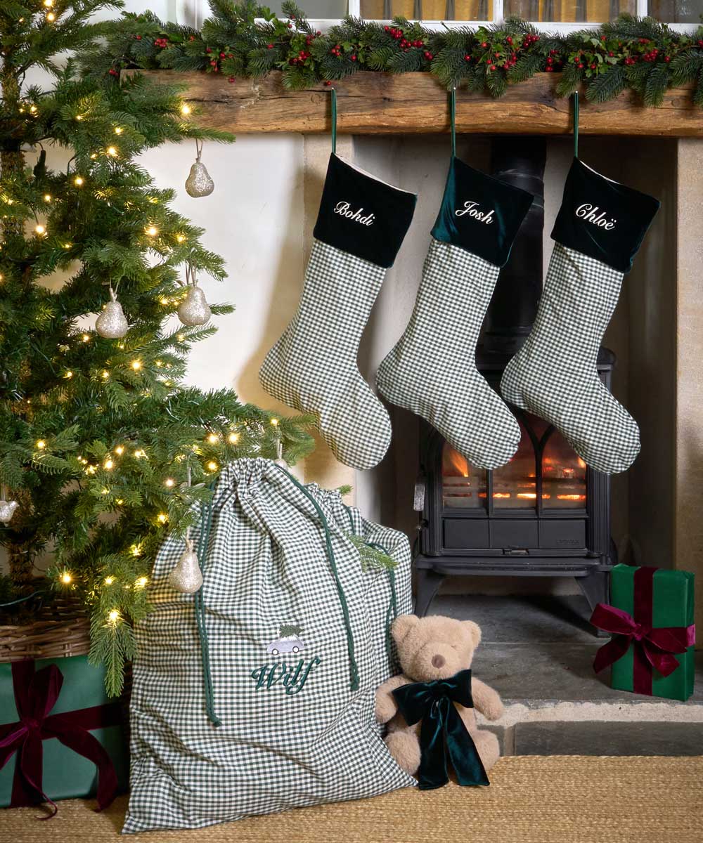 Three personalised Christmas stockings with green and white gingham check patterns and forest green velvet cuffs hang on a rustic wooden mantel above a burning wood stove. A matching gingham gift bag with green drawstrings sits on the floor next to a teddy bear, with a decorated Christmas tree and wrapped presents nearby.