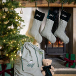 Three personalised Christmas stockings with green and white gingham check patterns and forest green velvet cuffs hang on a rustic wooden mantel above a burning wood stove. A matching gingham gift bag with green drawstrings sits on the floor next to a teddy bear, with a decorated Christmas tree and wrapped presents nearby.