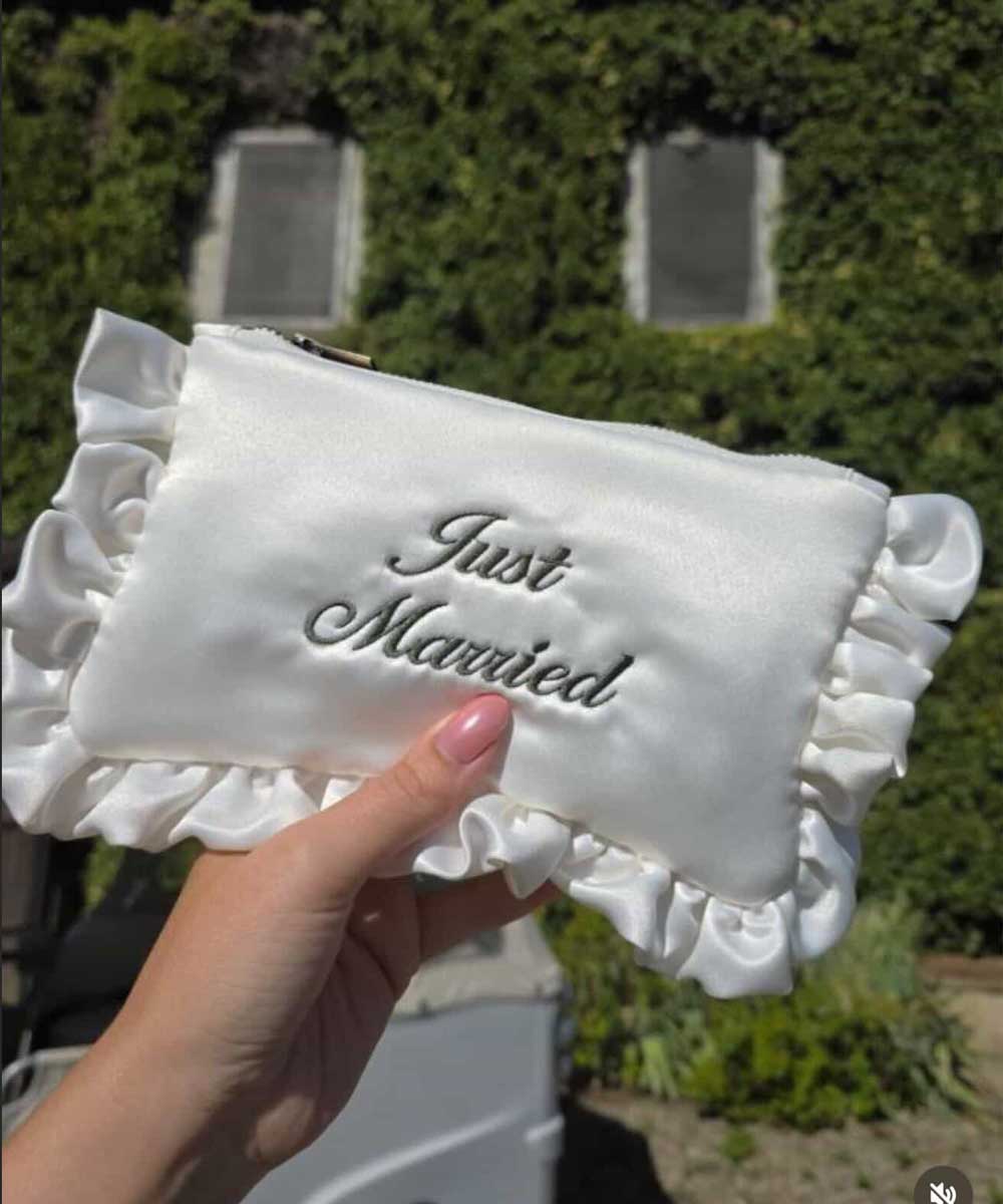 Ivory satin ruffled pouch with 'Just Married' script font embroidery held by a hand against a blurred outdoor background