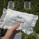 Ivory satin ruffled pouch with 'Just Married' script font embroidery held by a hand against a blurred outdoor background