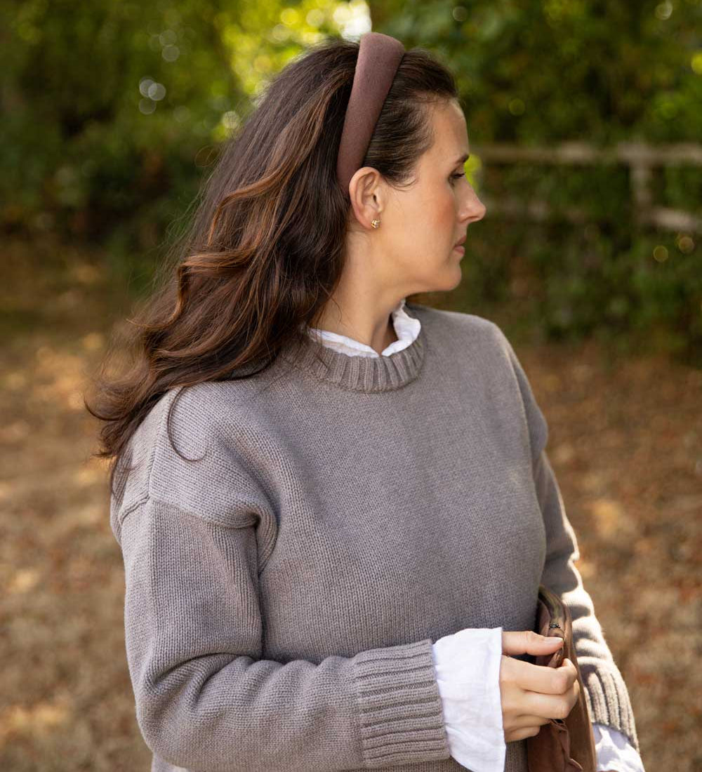 A woman in profile wearing a taupe gray knit sweater over a white collared shirt, accessorized with a brown headband, set against a soft-focus outdoor background of green foliage and brown ground.