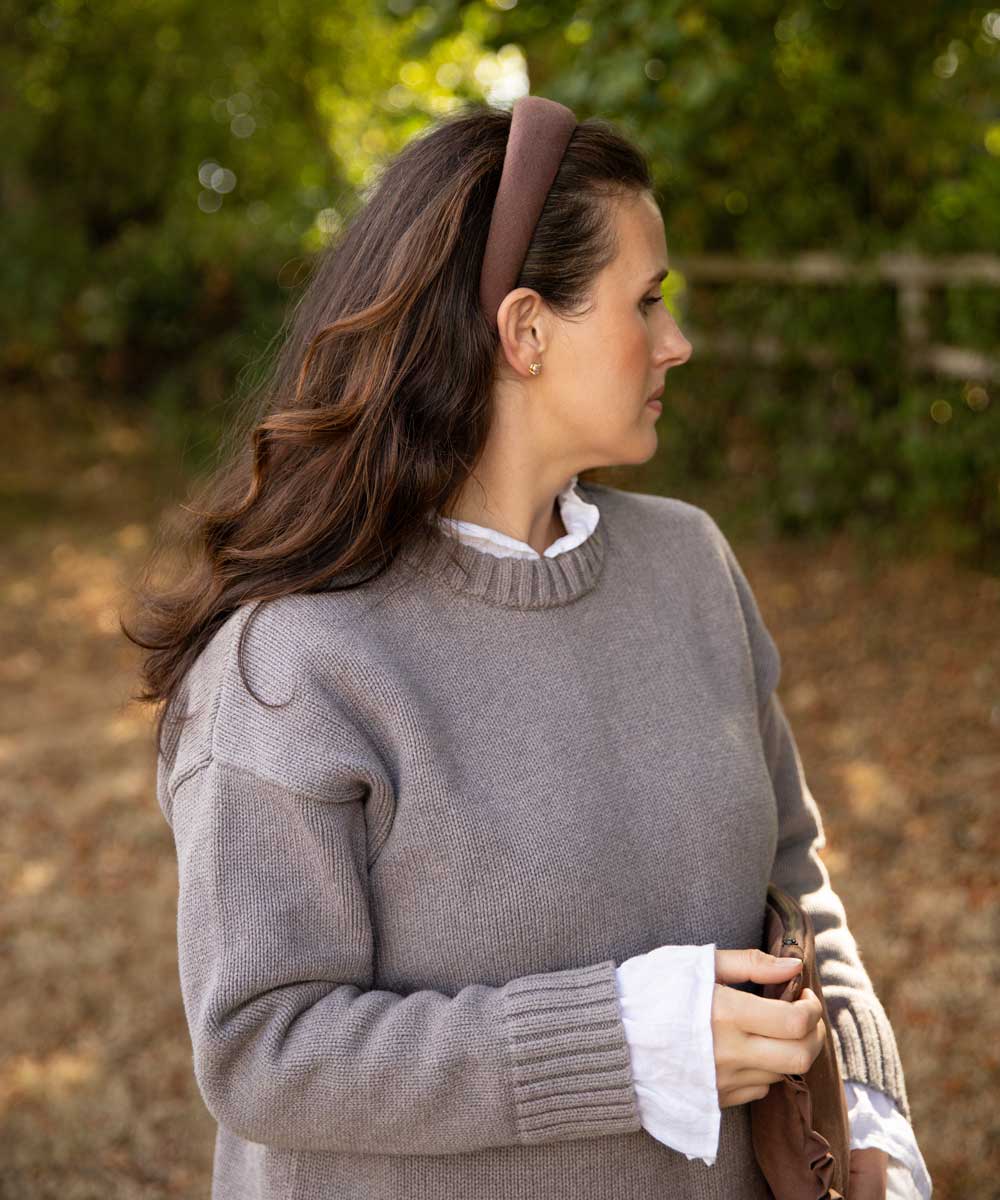 A woman in profile wearing a taupe gray knit sweater over a white collared shirt, accessorized with a brown headband, set against a soft-focus outdoor background of green foliage and brown ground.