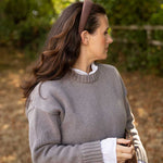 A woman in profile wearing a taupe gray knit sweater over a white collared shirt, accessorized with a brown headband, set against a soft-focus outdoor background of green foliage and brown ground.