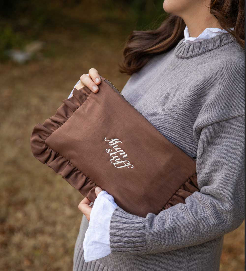 A woman in a gray knit sweater holds a brown ruffled clutch bag with white embroidered text reading 'Mum stuff' in an outdoor setting.