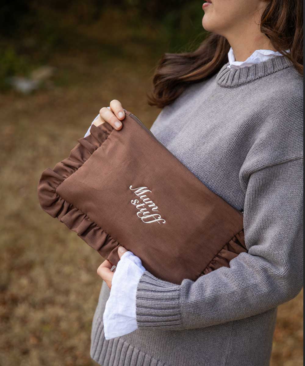 A woman in a gray knit sweater holds a brown ruffled clutch bag with white embroidered text reading 'Mum stuff' in an outdoor setting.