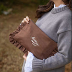 A woman in a gray knit sweater holds a brown ruffled clutch bag with white embroidered text reading 'Mum stuff' in an outdoor setting.