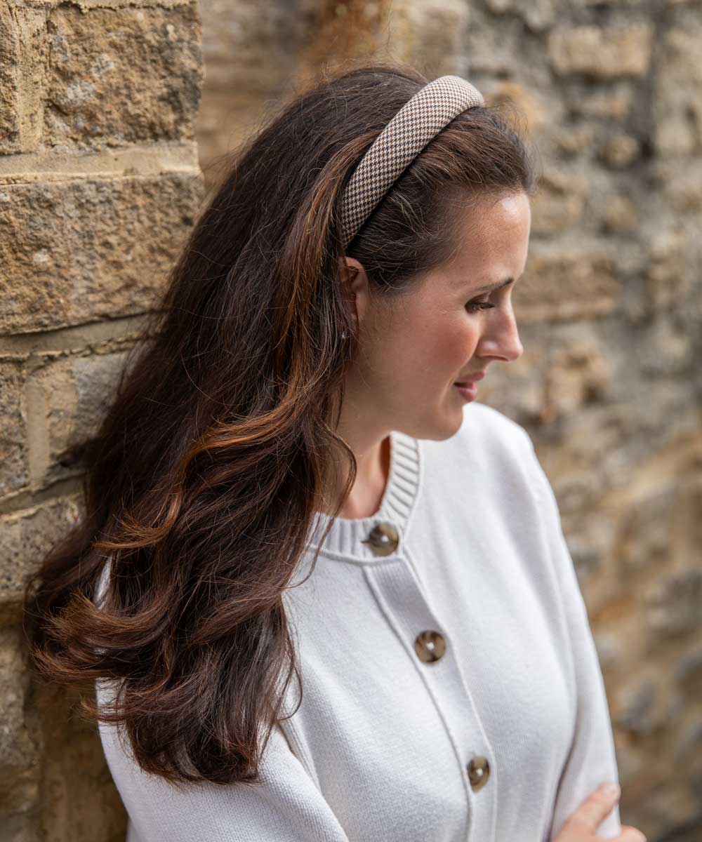 A woman with long brown hair wearing a beige and brown checkered headband and a white cardigan, standing in profile against a rustic stone wall.