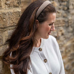 A woman with long brown hair wearing a beige and brown checkered headband and a white cardigan, standing in profile against a rustic stone wall.