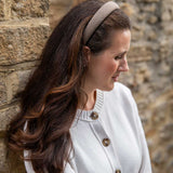 A woman with long brown hair wearing a beige and brown checkered headband and a white cardigan, standing in profile against a rustic stone wall.