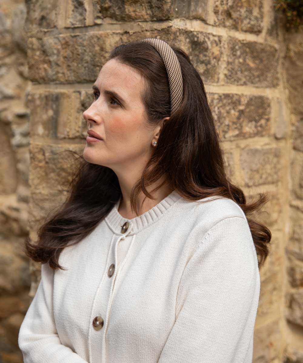 A woman with long dark brown hair wearing a beige and brown textured headband and a cream-colored knit cardigan, standing in front of a rustic stone wall.