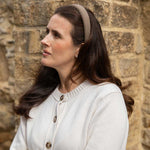 A woman with long dark brown hair wearing a beige and brown textured headband and a cream-colored knit cardigan, standing in front of a rustic stone wall.