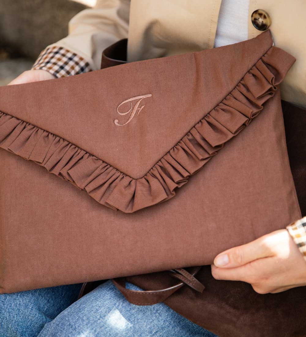 A person holds a brown envelope-style clutch bag with a ruffled flap and an embroidered monogram. The clutch is made of a textured fabric and shown in a close-up lifestyle shot.