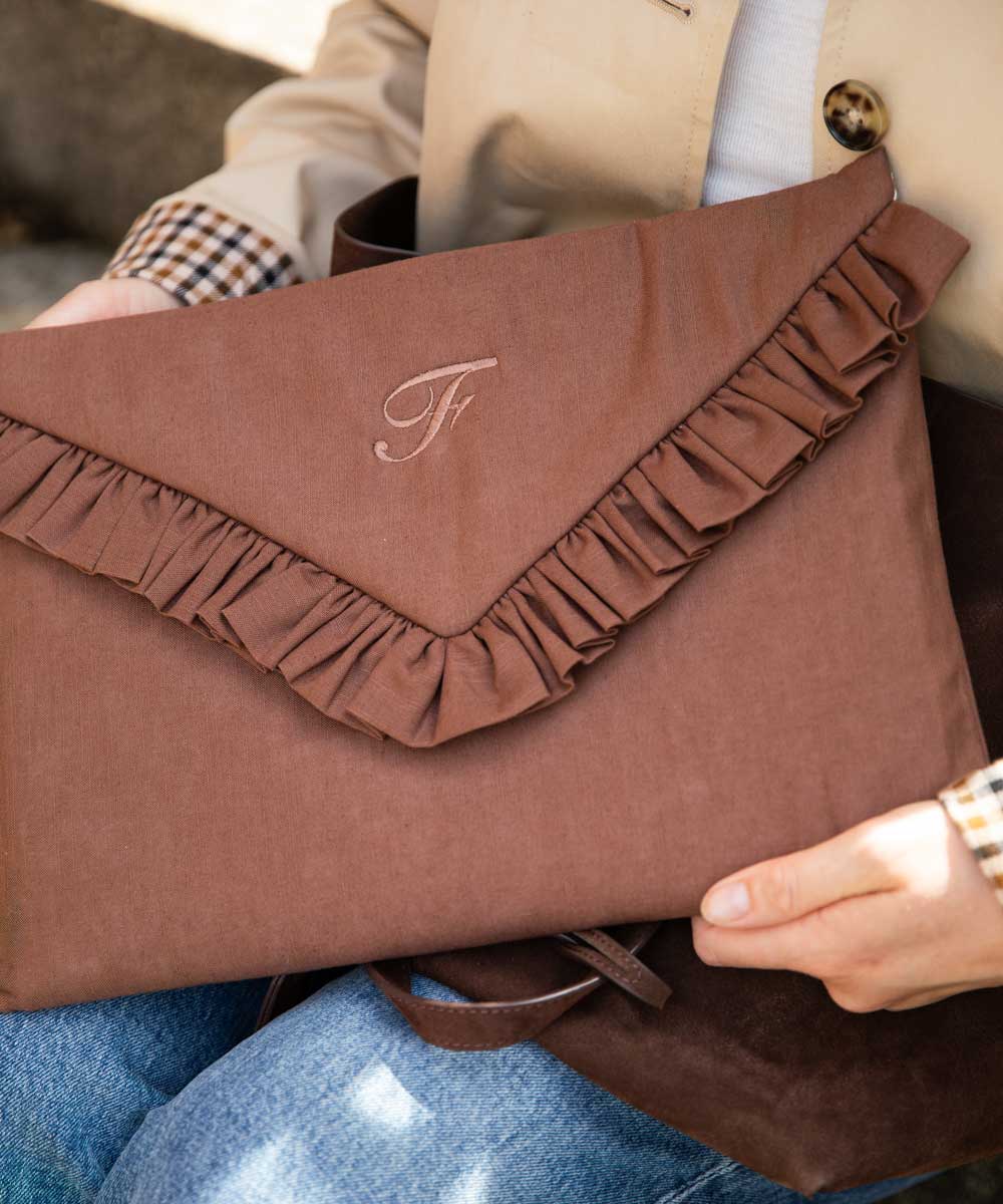 A person holds a brown envelope-style clutch bag with a ruffled flap and an embroidered monogram. The clutch is made of a textured fabric and shown in a close-up lifestyle shot.