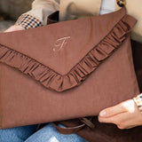 A person holds a brown envelope-style clutch bag with a ruffled flap and an embroidered monogram. The clutch is made of a textured fabric and shown in a close-up lifestyle shot.