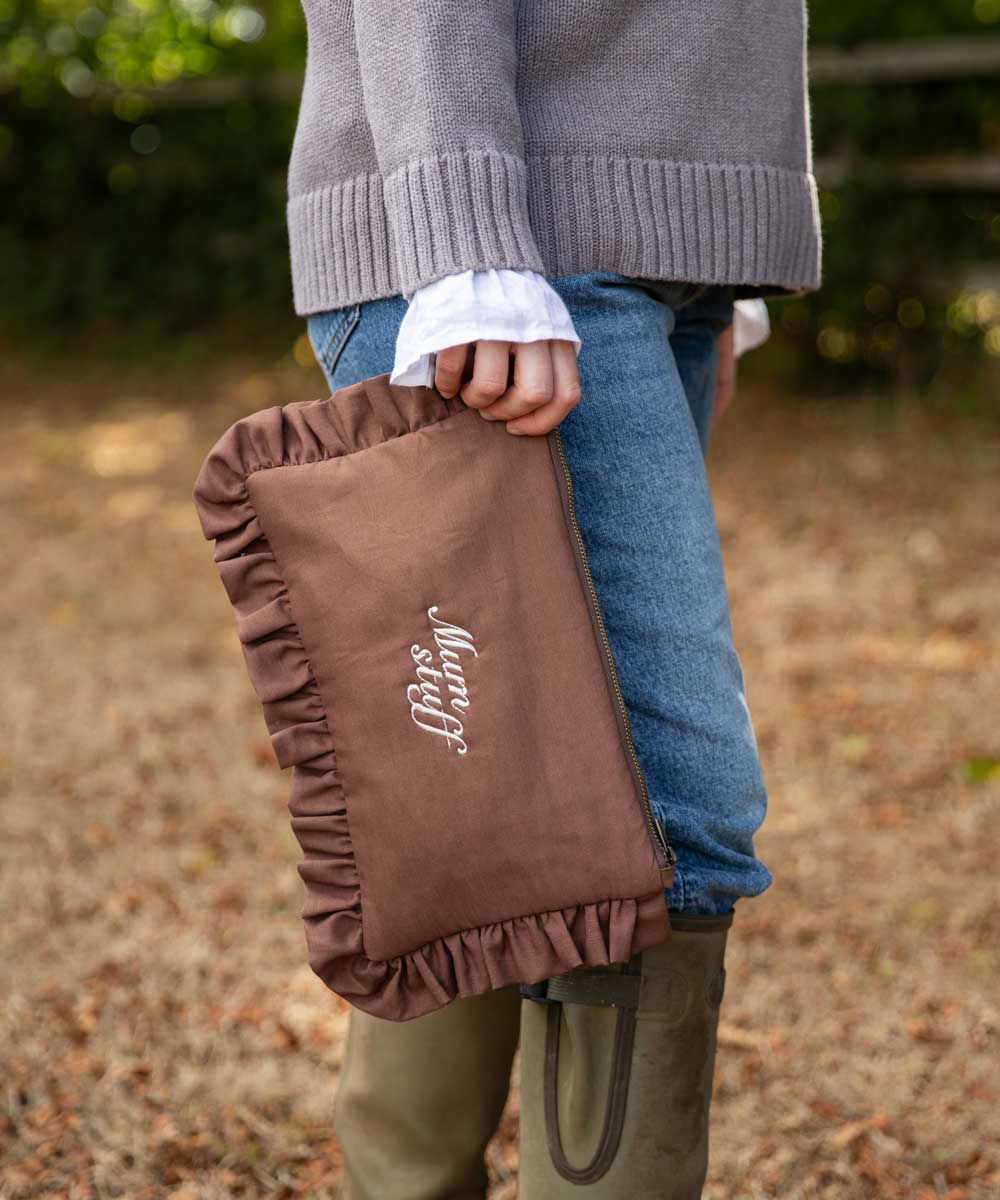 A person outdoors holding a brown ruffled fabric bag with white script embroidery that reads 'mum stuff'. The person is wearing a gray knit sweater, blue jeans, and olive green rain boots, with a blurred natural background.