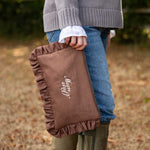 A person outdoors holding a brown ruffled fabric bag with white script embroidery that reads 'mum stuff'. The person is wearing a gray knit sweater, blue jeans, and olive green rain boots, with a blurred natural background.