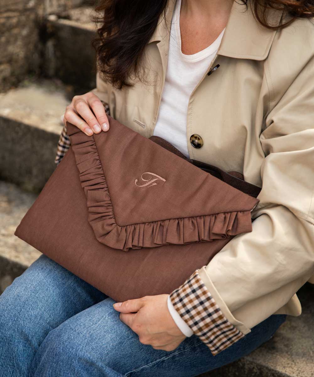A woman in a beige trench coat and blue jeans holds a brown ruffled laptop case bag with a monogram logo.