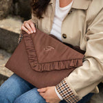 A woman in a beige trench coat and blue jeans holds a brown ruffled laptop case bag with a monogram logo.