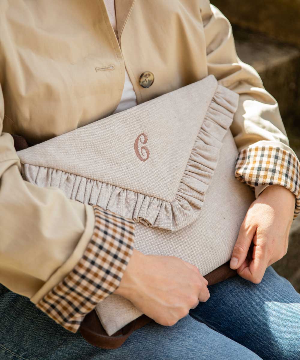 A beige fabric  laptop case with a ruffled envelope-style flap and a monogrammed 'G' on the front, held by a person wearing a beige jacket and plaid shirt.
