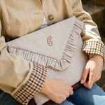 A beige fabric  laptop case with a ruffled envelope-style flap and a monogrammed 'G' on the front, held by a person wearing a beige jacket and plaid shirt.