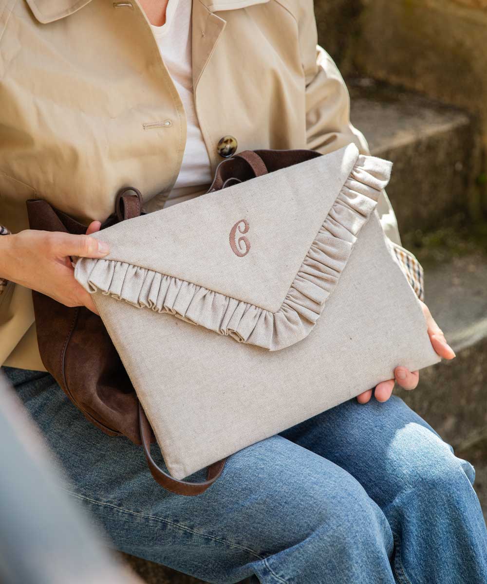 A person in a beige trench coat and blue jeans holds a light beige linen envelope-style laptop case with a ruffled flap and a monogrammed 'G' on the front. A dark brown leather bag is tucked under the coat.