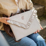 A person in a beige trench coat and blue jeans holds a light beige linen envelope-style laptop case with a ruffled flap and a monogrammed 'G' on the front. A dark brown leather bag is tucked under the coat.