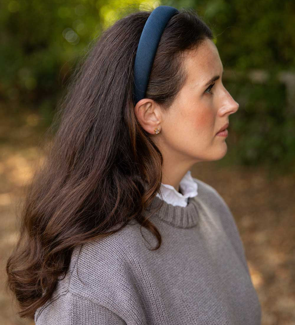 A woman in profile wearing a navy blue fabric headband, a gray knit sweater, and a white ruffled collar, set against a soft-focus outdoor background of green foliage.