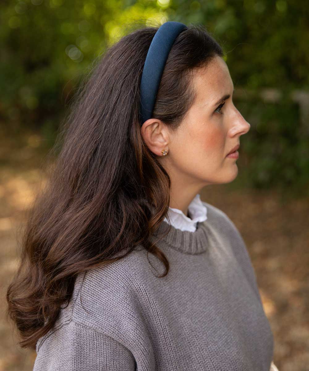 A woman in profile wearing a navy blue fabric headband, a gray knit sweater, and a white ruffled collar, set against a soft-focus outdoor background of green foliage.