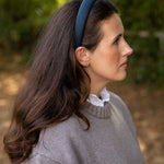 A woman in profile wearing a navy blue fabric headband, a gray knit sweater, and a white ruffled collar, set against a soft-focus outdoor background of green foliage.