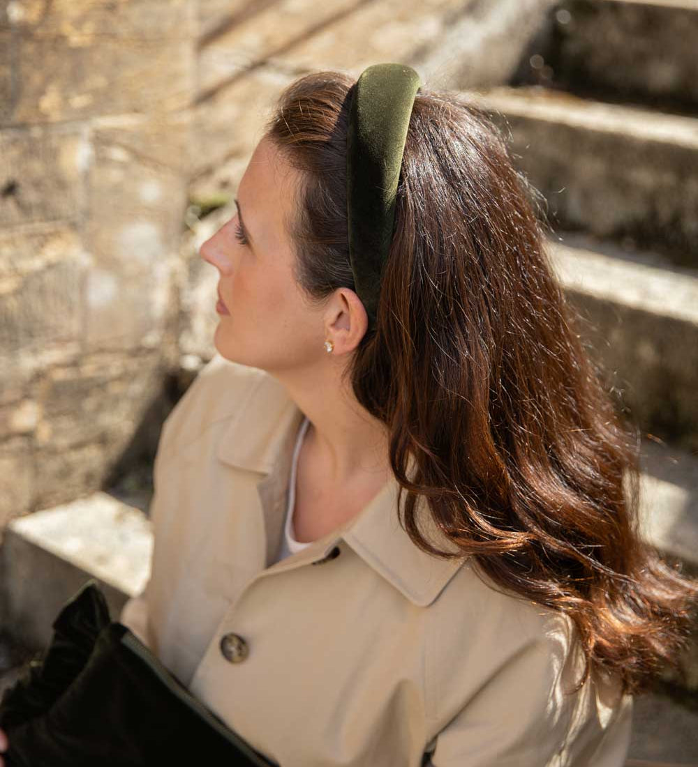 A woman with long brown hair wearing an olive green velvet headband, styled with a beige trench coat, standing on stone steps.