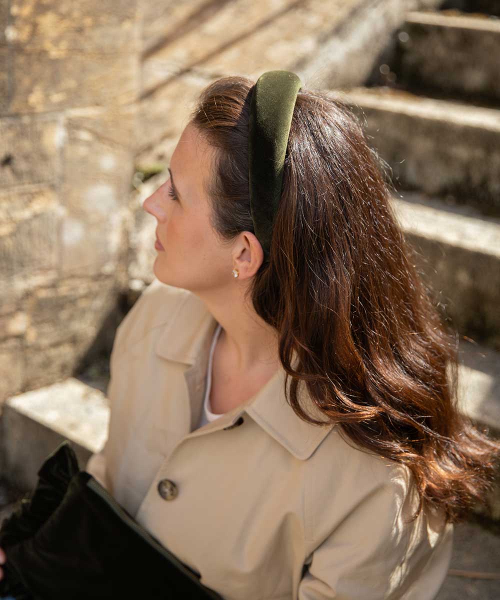 A woman with long brown hair wearing an olive green velvet headband, styled with a beige trench coat, standing on stone steps.