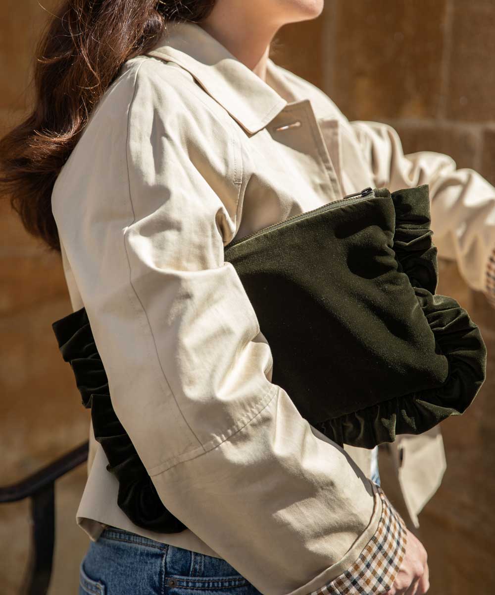 A person wearing a light beige trench-style jacket and blue jeans holds a dark olive green pleated clutch bag against their arm.