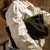 A person wearing a light beige trench-style jacket and blue jeans holds a dark olive green pleated clutch bag against their arm.