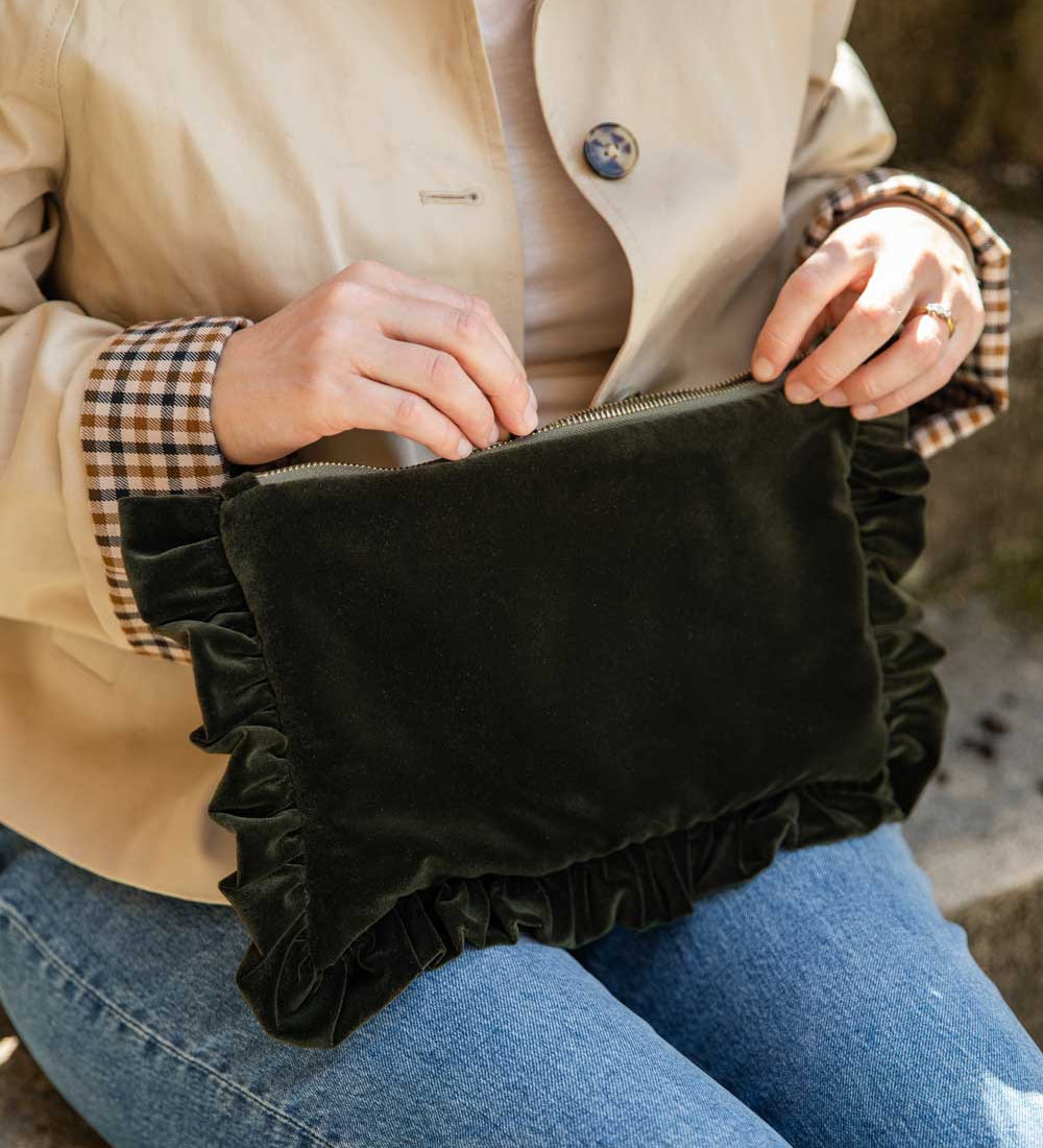 A person wearing a beige trench coat and blue jeans holds a olive velvet clutch bag with ruffled trim in their lap.