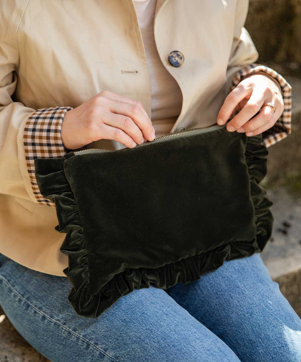 A person wearing a beige trench coat and blue jeans holds a olive velvet clutch bag with ruffled trim in their lap.