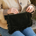 A person wearing a beige trench coat and blue jeans holds a olive velvet clutch bag with ruffled trim in their lap.